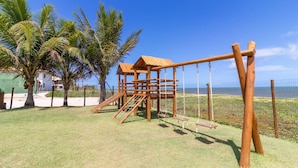 A wooden playground with slides and swings, surrounded by palm trees and a clear blue sky. - IT27 Village 3 Suítes Pé na Areia (Lauro de Freitas)
