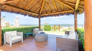 A covered outdoor seating area with wicker furniture, a wooden deck, and a view of the beach. - IT27 Village 3 Suítes Pé na Areia (Lauro de Freitas)