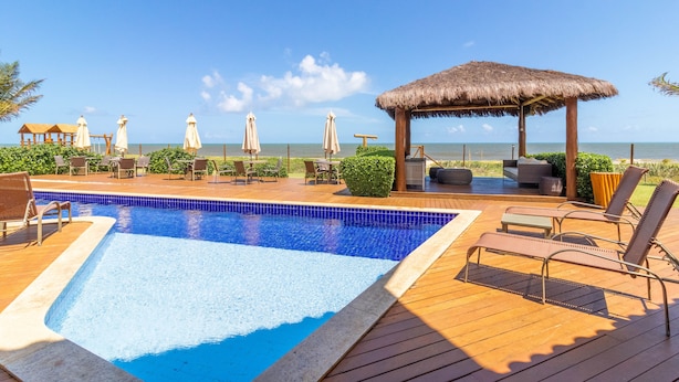 A pool area with lounge chairs, a thatched-roof gazebo, and a view of the ocean. - IT27 Village 3 Suítes Pé na Areia (Lauro de Freitas)