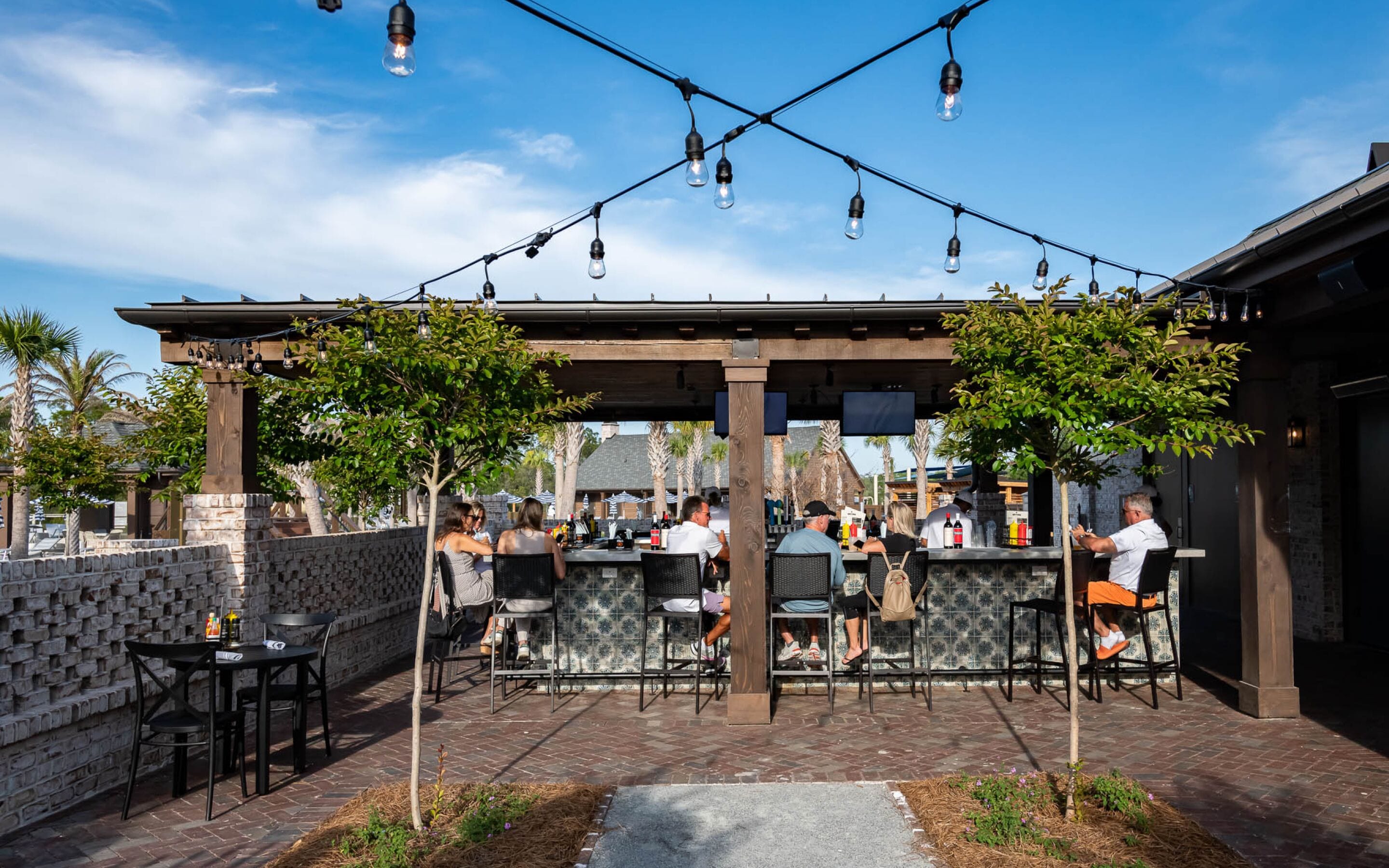 An outdoor patio area with tables and chairs, a bar with a counter, and string lights overhead.