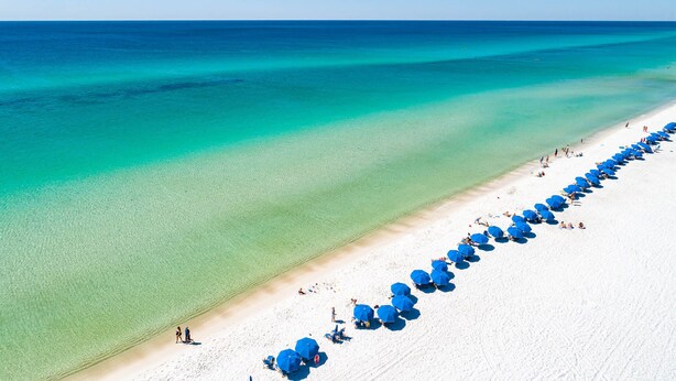 An aerial view of a beach with a long row of blue umbrellas and people on the sand. - Camp Creek Inn (Panama City Beach)