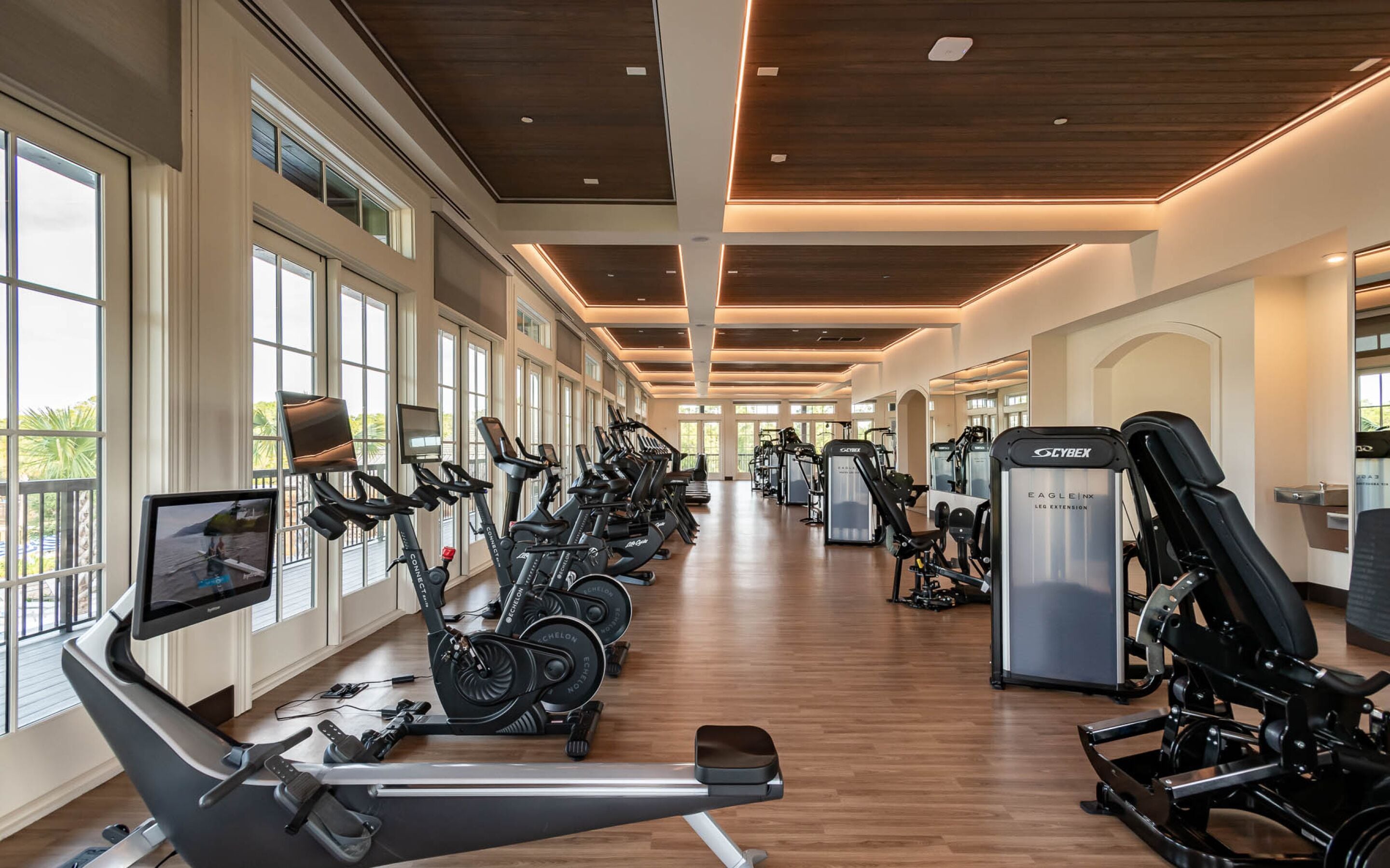 A gym with multiple exercise machines, large windows, and a wooden floor.