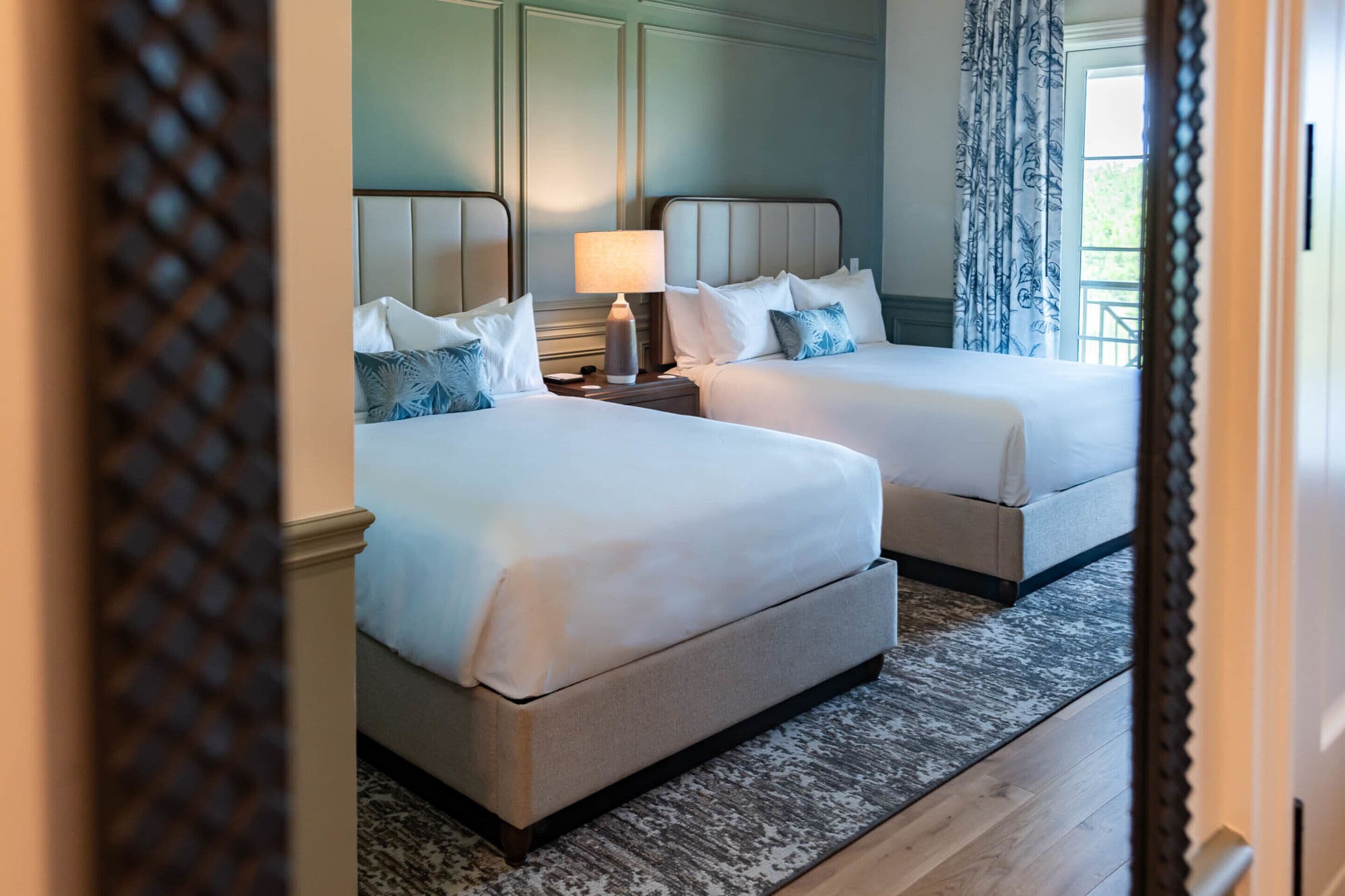 Quarto de hotel com duas camas, uma mesa de cabeceira com abajur e vista para o exterior através de uma janela com cortinas.