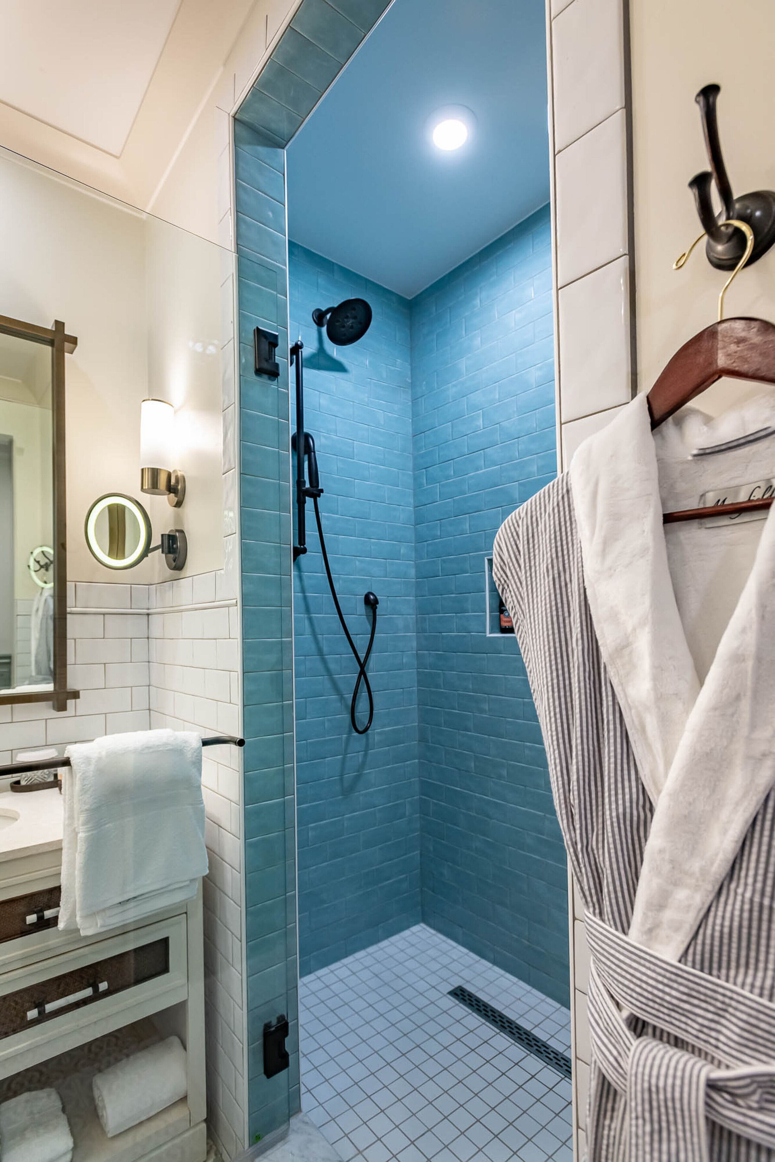 Une salle de bain moderne avec une douche dont le carrelage est bleu, un lavabo blanc avec un miroir et un porte-serviettes avec un peignoir.