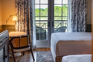 Une chambre d’hôtel avec un lit, un bureau doté d’une lampe, un fauteuil en osier et une vue sur l’extérieur grâce à une porte vitrée.