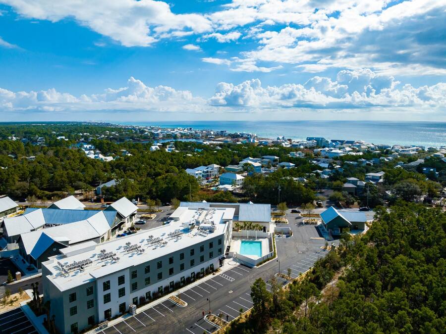 An aerial view of a coastal town with a large building featuring a rooftop pool and parking lot.