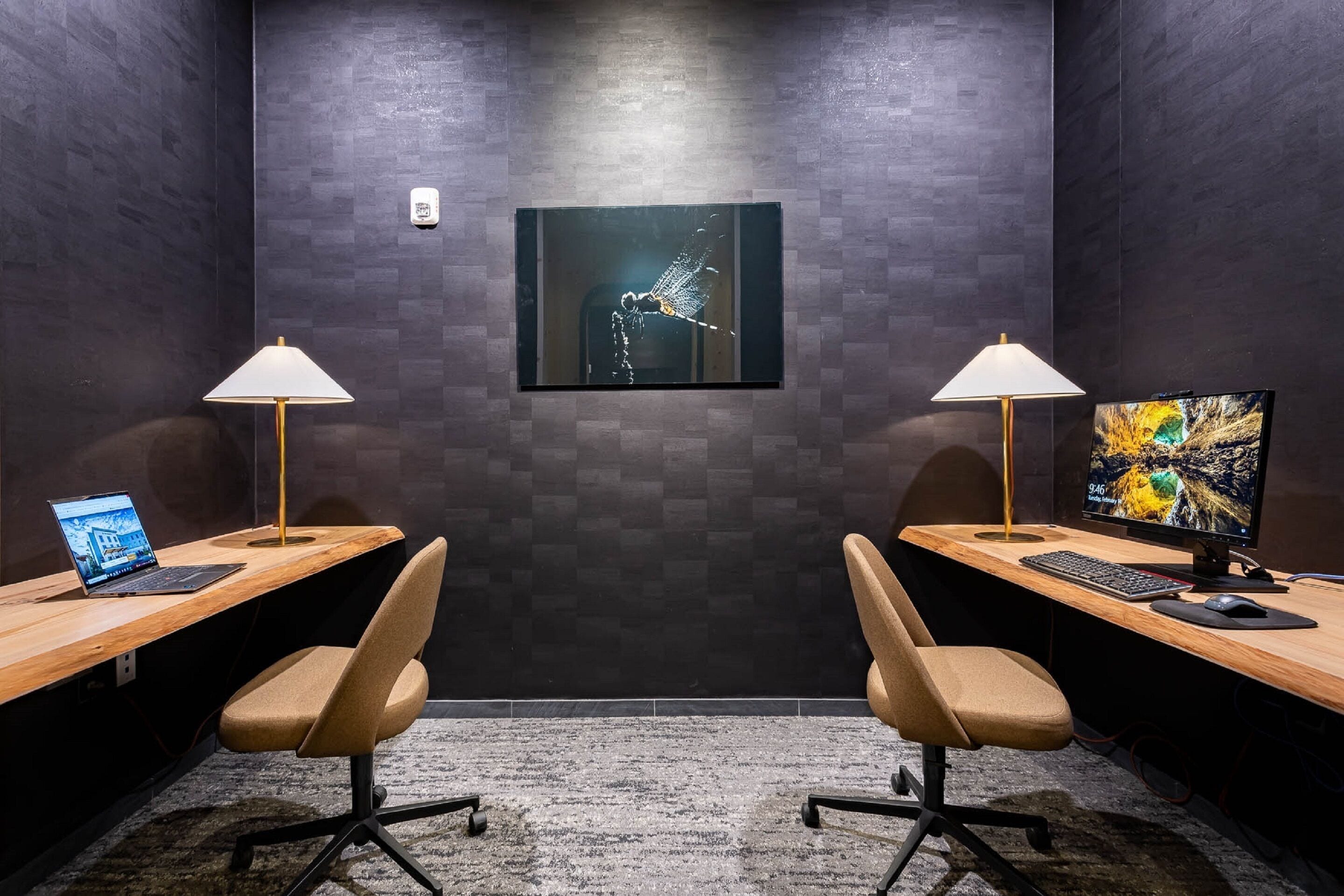 A conference room with two desks, each with a laptop, a keyboard, and a monitor. There are two chairs, a wall-mounted lamp, and a framed artwork on the wall.