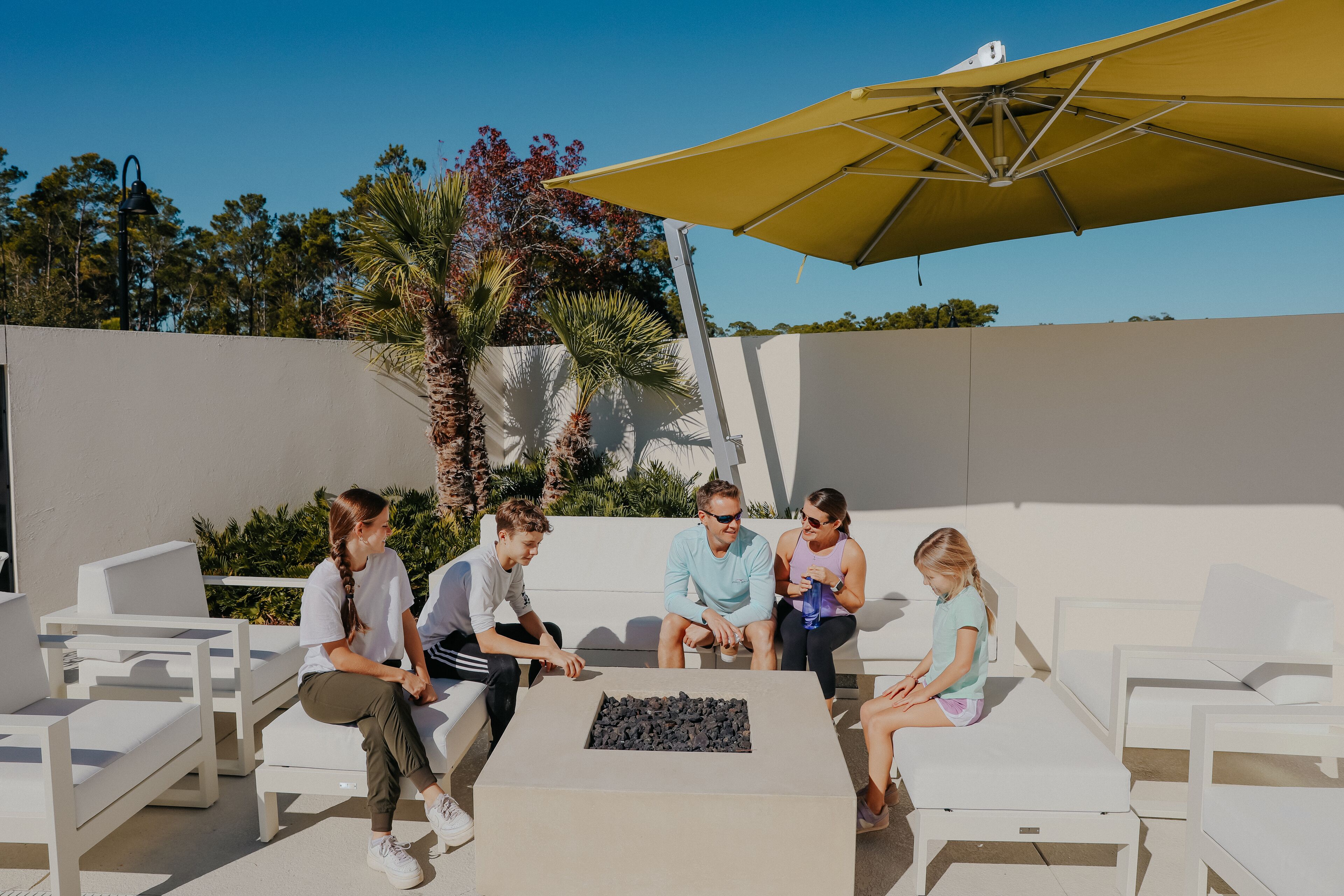 A group of people seated around a table with a fire feature, under a large umbrella, in an outdoor setting with white furniture and palm trees.