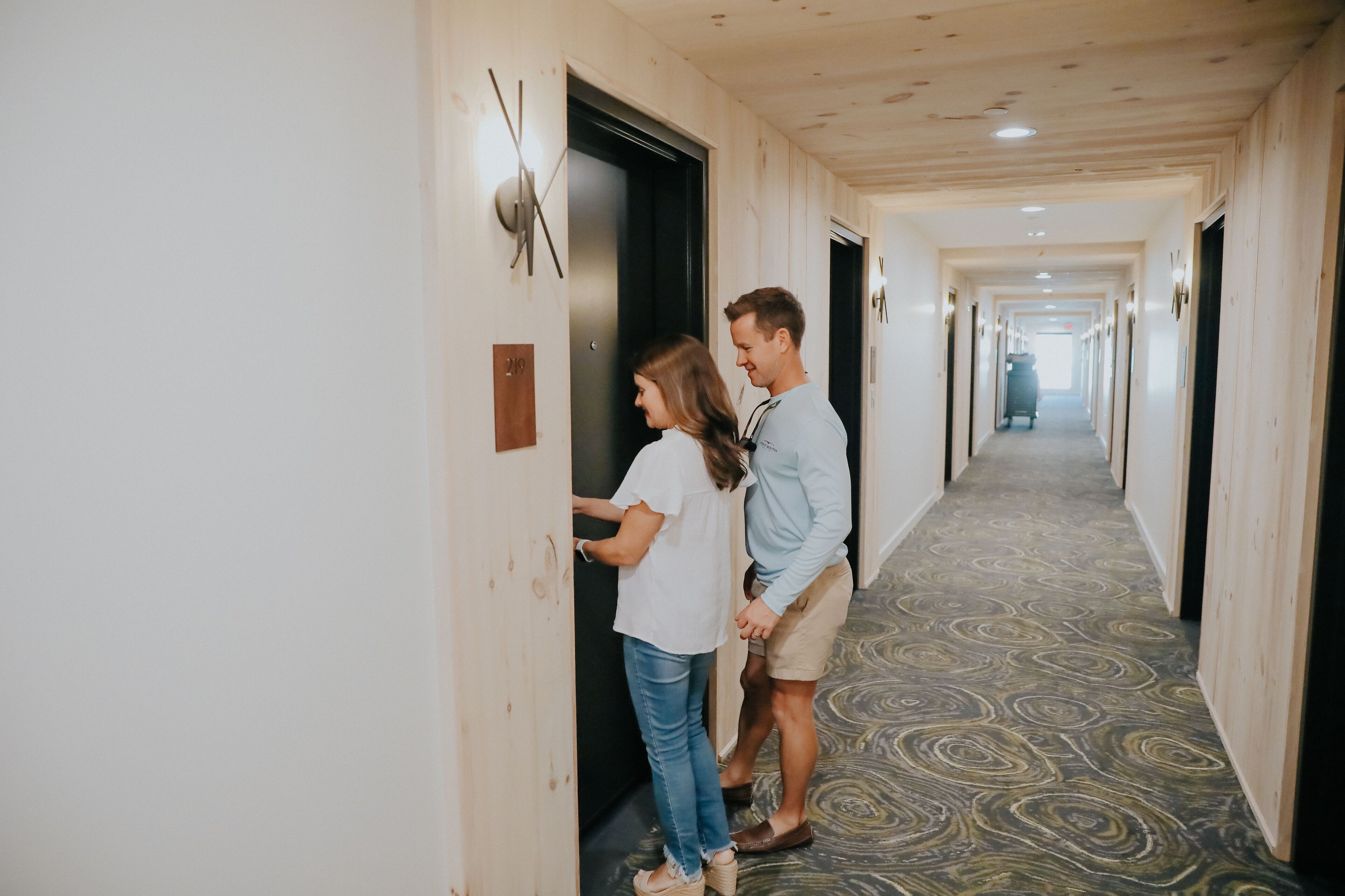 A hotel hallway with a patterned carpet, wooden walls, and a door with a handle.