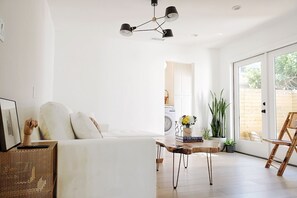 A bright, modern living room with a sofa, a coffee table, and a dining area. There is a large window providing natural light and a view of the outside.
