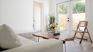 A bright living room with a white sofa, a wooden coffee table with books and flowers, a wicker chair, and a sliding glass door leading to an outdoor area.