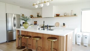 A modern kitchen with a central island, bar stools, a stainless steel refrigerator, and a hanging light fixture.