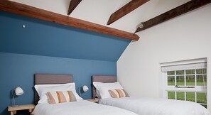 A bedroom with two beds, wooden beams, a window with blinds, and a view of greenery outside.
