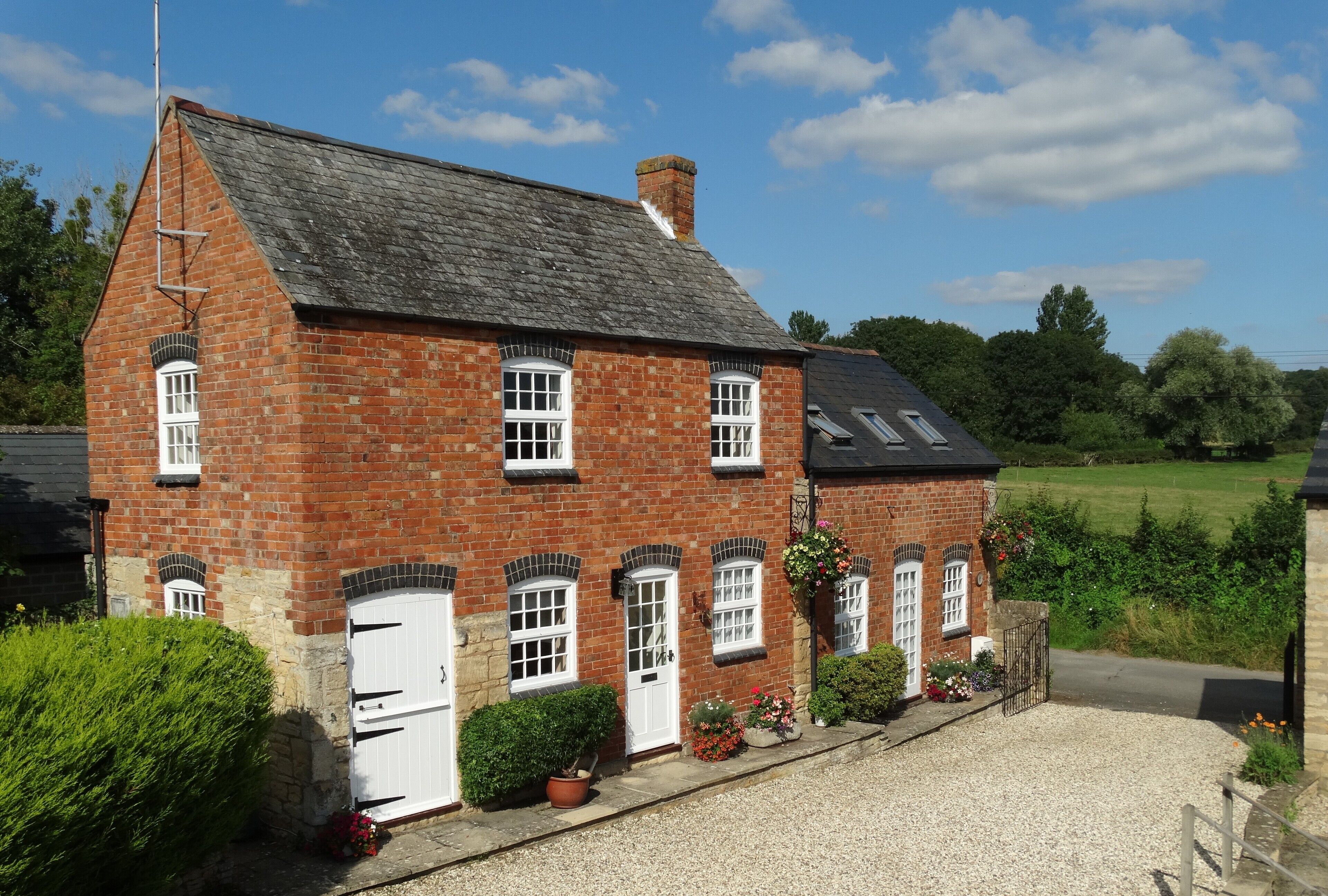 A red brick house with white doors and windows, a gravel driveway, and a garden with potted plants and flowers.
