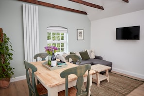 A living room with a wooden dining table, chairs, a sofa, a TV mounted on the wall, a window with blinds, and a potted plant.