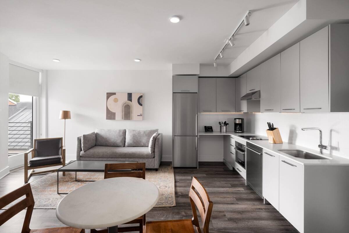 A modern kitchen with grey cabinets, a stainless steel refrigerator, and a built-in oven. There is a dining area with a round table and chairs, and a grey sofa in the living room area.