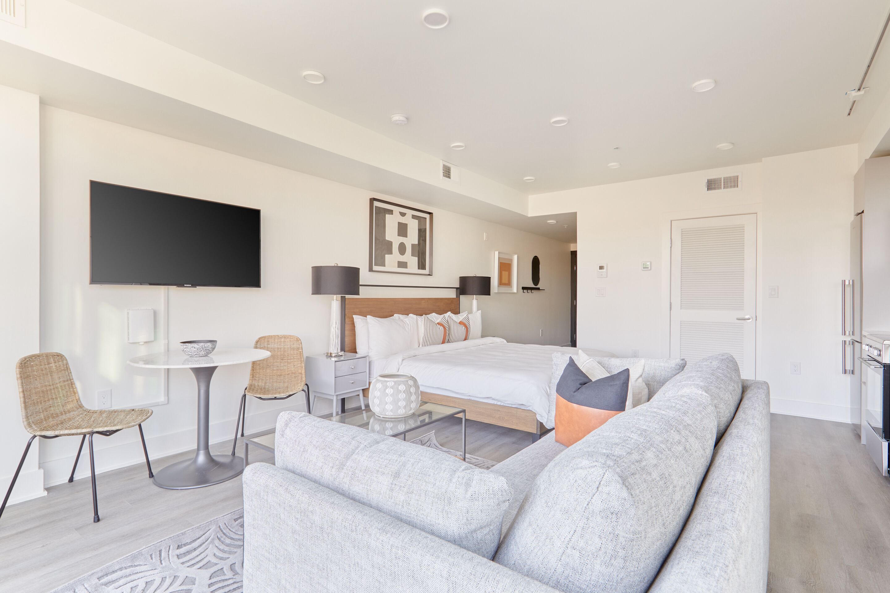 A modern hotel room with a large bed, a sofa, a TV, and a dining area with chairs and a table.