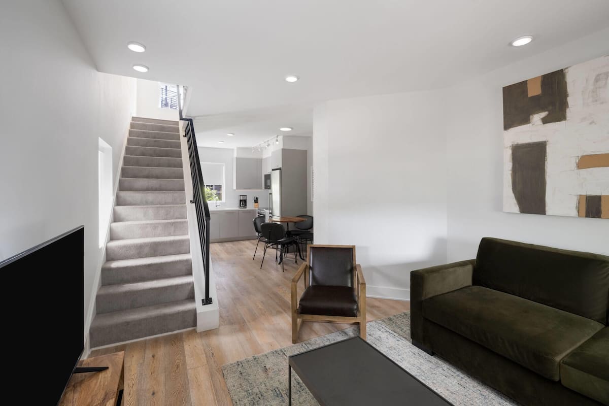 A modern living room with a staircase, a sofa, a coffee table, and a dining area in the background.