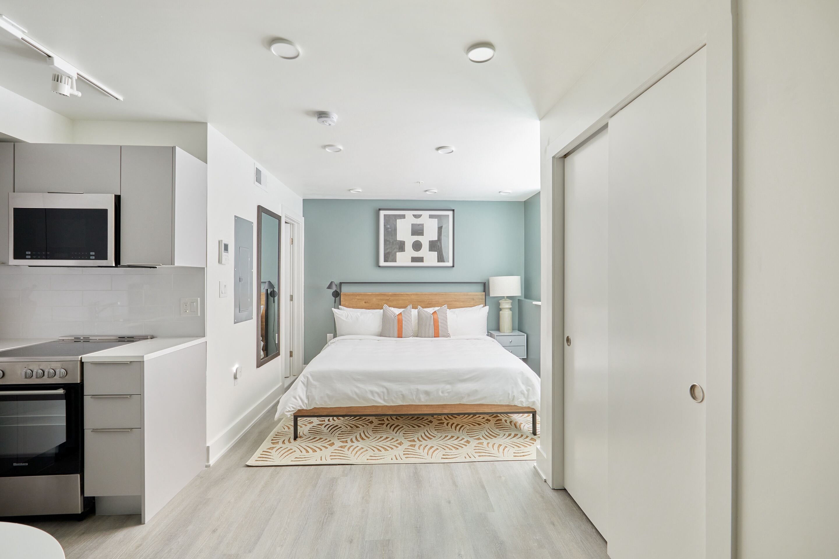 A modern bedroom with a bed, bedside table, and a view of the kitchen.