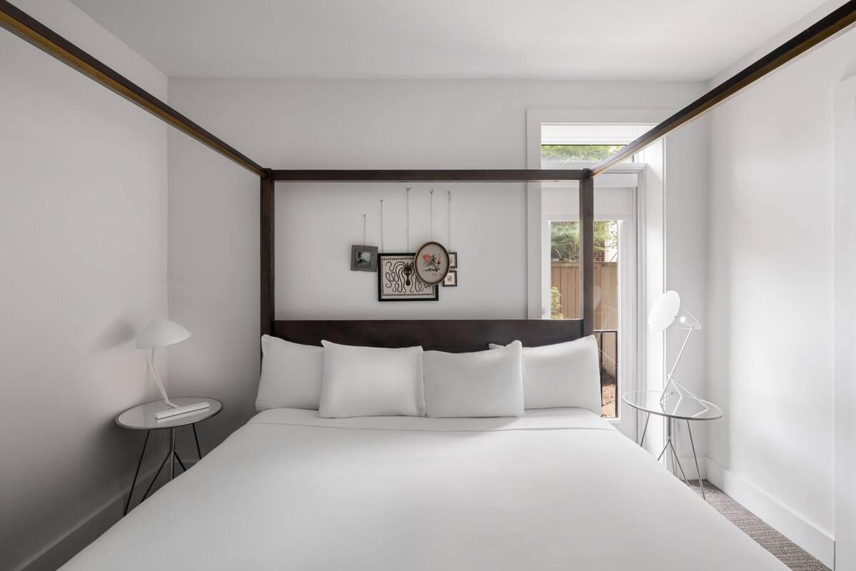 A modern bedroom with a large bed, a bedside table with a lamp, a small side table, and a wall-mounted artwork.