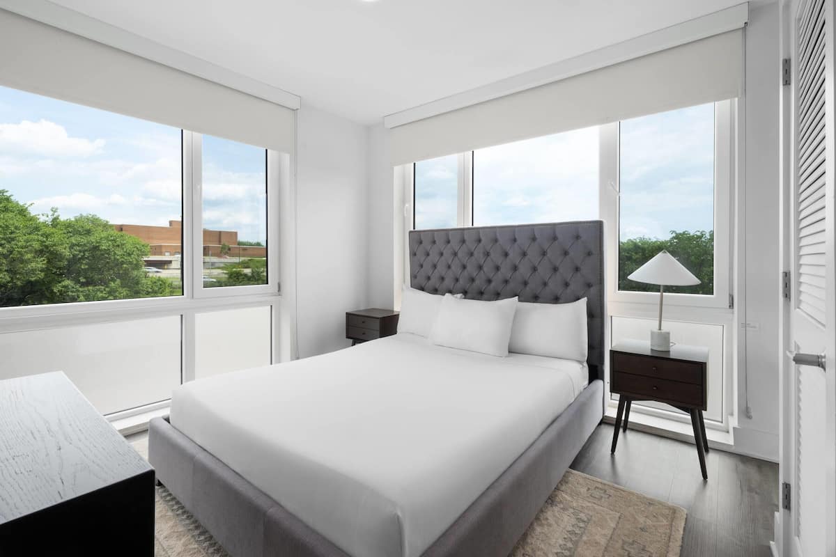 A modern bedroom with a large bed, a nightstand, a lamp, and a view of the outdoors.