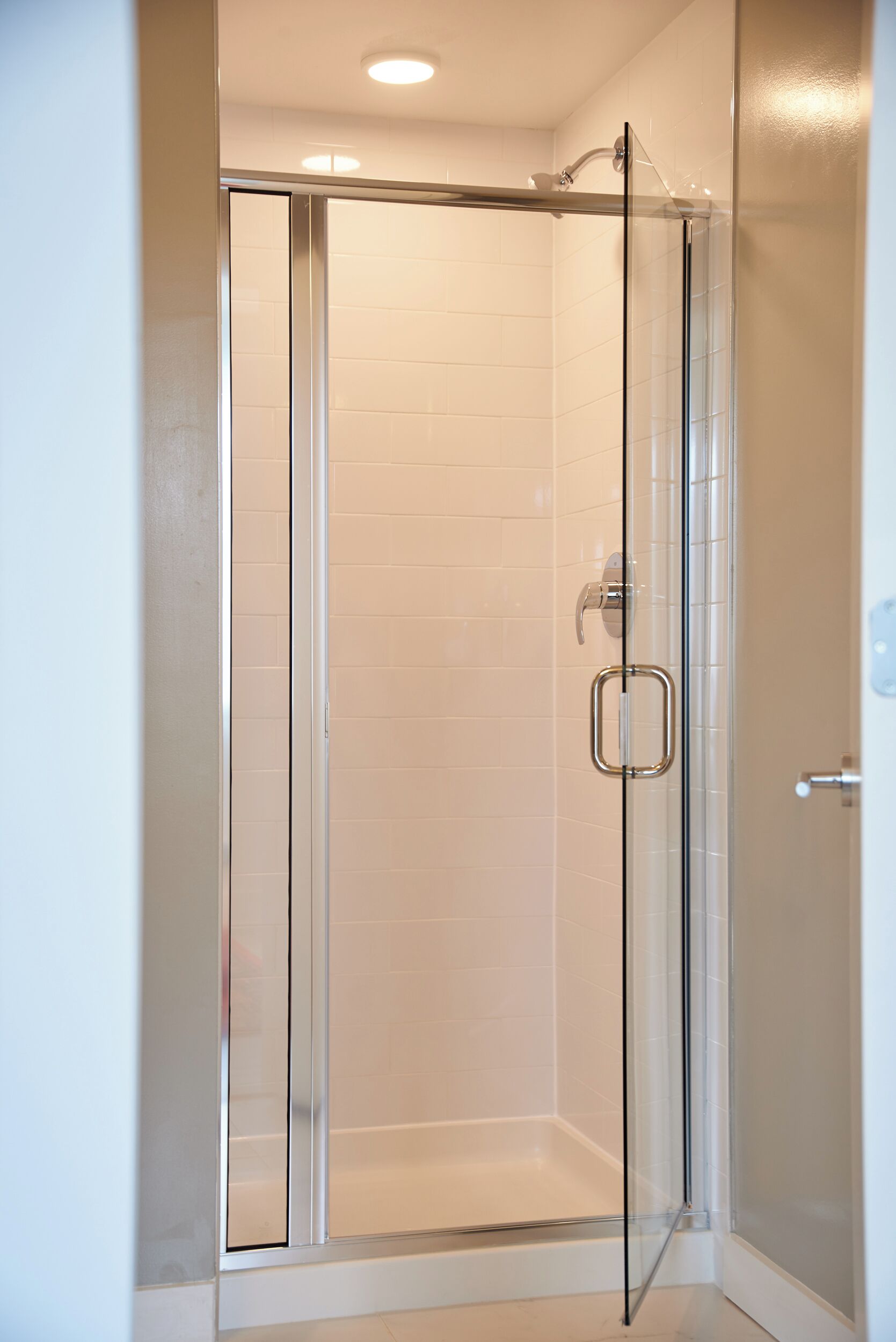 Une douche fermée par des parois de verre, avec un mur recouvert de carreaux blancs, une porte en verre transparent et une poignée en métal.