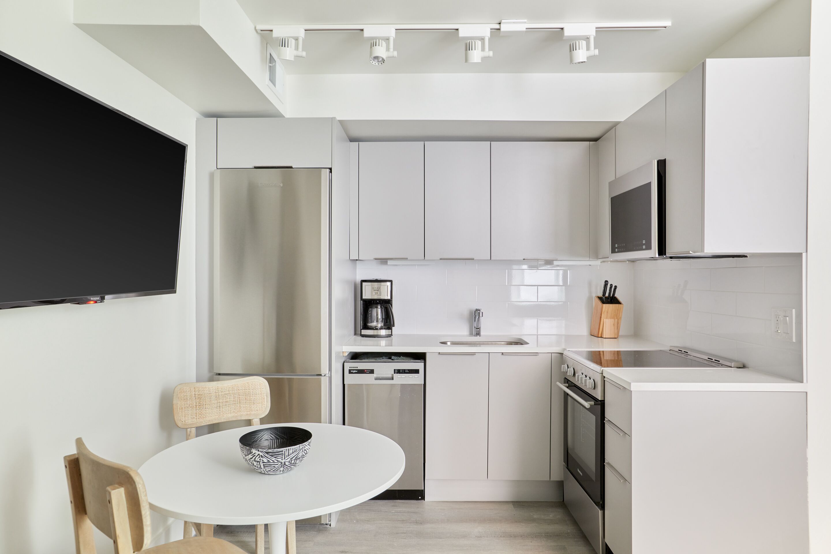 Una cocina moderna con armarios blancos, un refrigerador de acero inoxidable, un lavavajillas, un microondas y una estufa. Hay una mesa de comedor con dos sillas y un bol sobre ella.