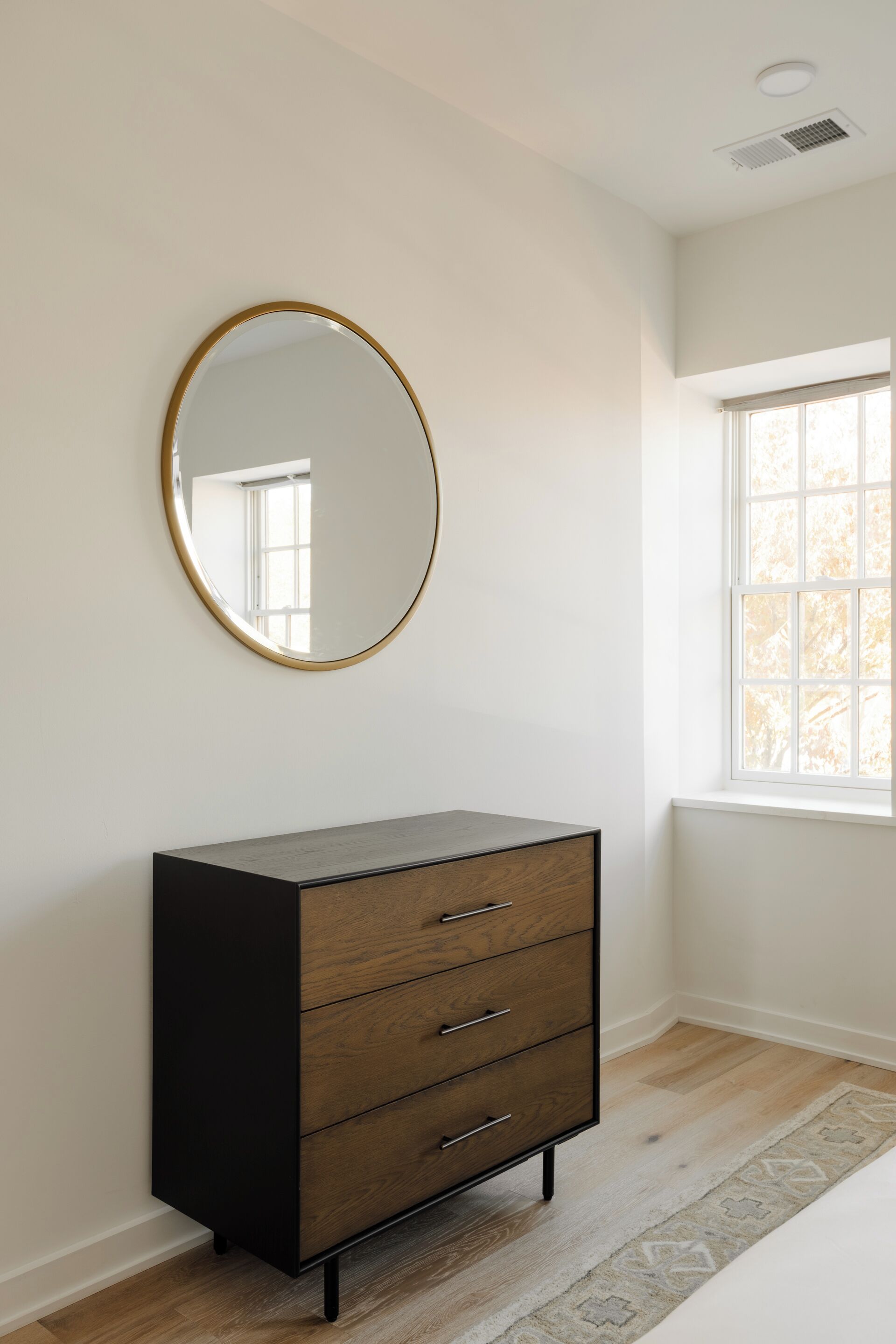A room with a wooden dresser, a round mirror, and a window.