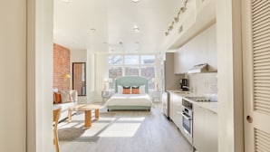 Una habitación de hotel moderna con una cama grande, una zona de estar con sofá y sillones, un área de cocina y vistas al exterior.