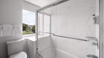 Un baño moderno con ducha envuelta en vidrio, paredes de mármol blanco, inodoro y ventana con vistas al exterior.