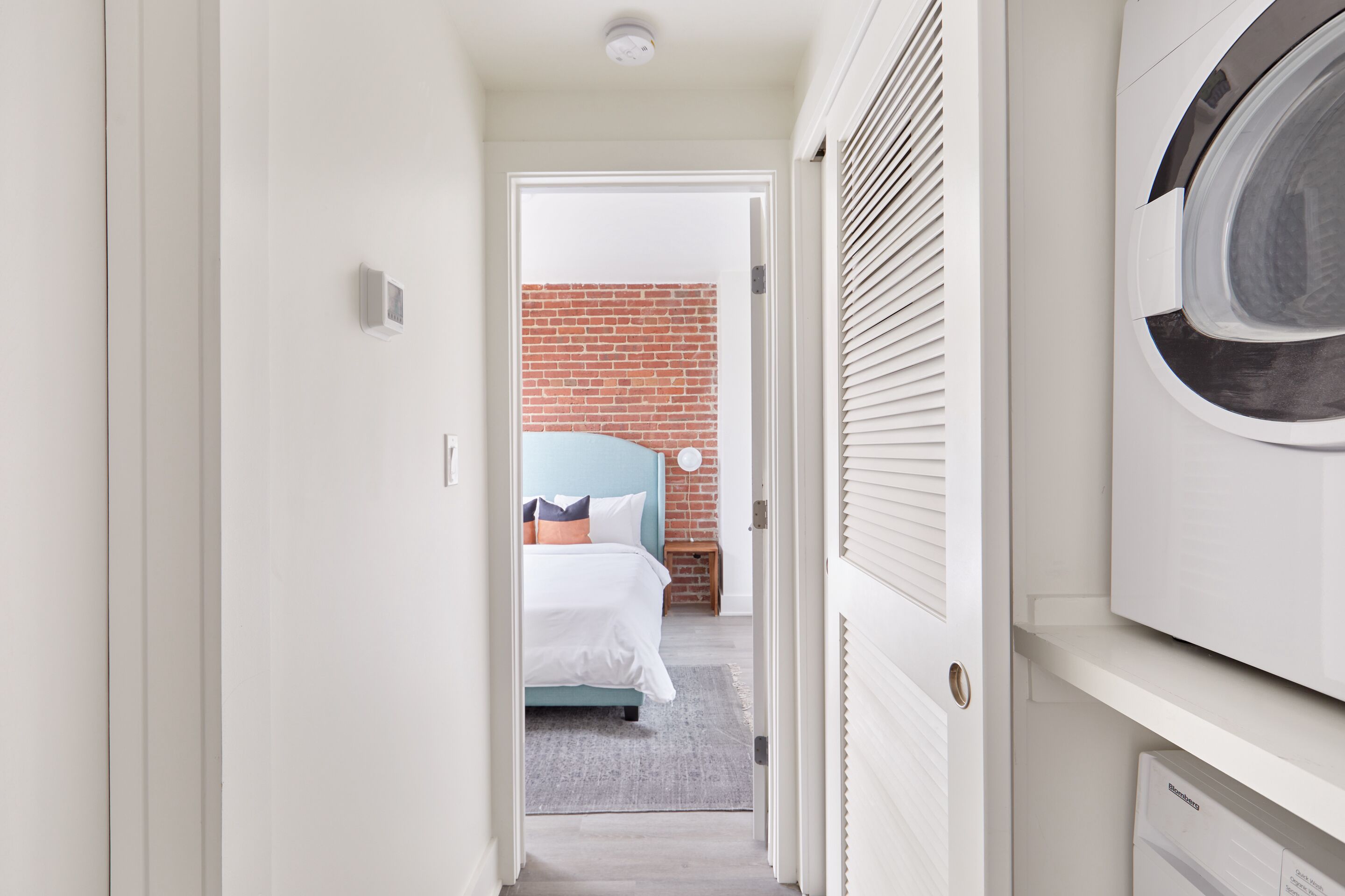 Un couloir étroit mène à une chambre dotée d’un mur en briques, d’un lit et d’un coin buanderie avec un lave-linge.