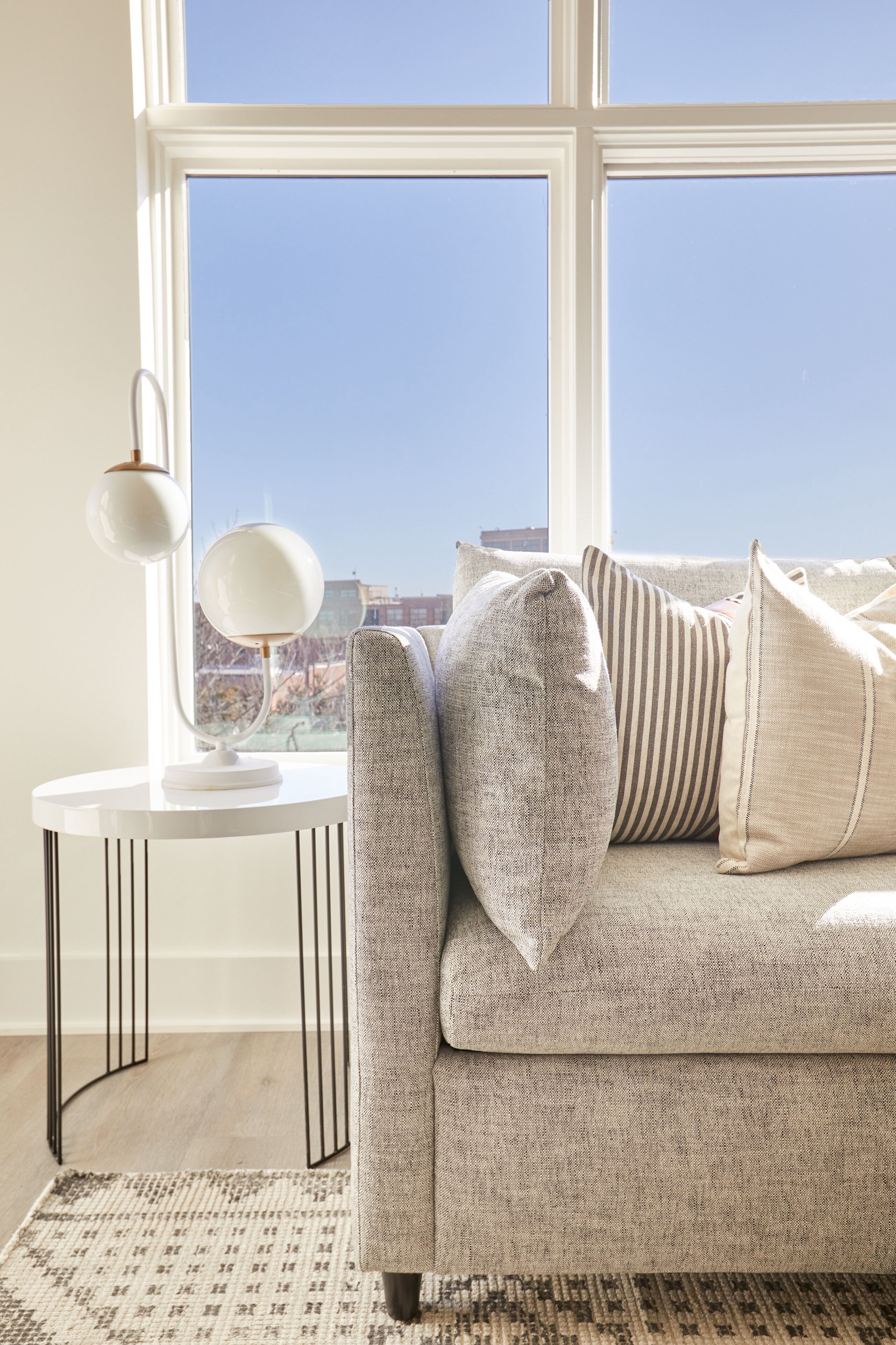 Una sala de estar moderna con un sofá gris, una mesa redonda con una lámpara blanca y vistas al exterior a través de grandes ventanales.