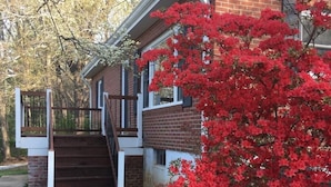 Une maison à deux étages, avec une façade en briques, un porche en bois et un arbuste à fleurs rouges.