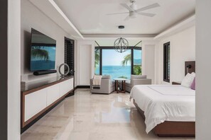 Quarto de hotel moderno com uma cama grande, televisão, área de estar com vista para o oceano e ventilador de teto.
