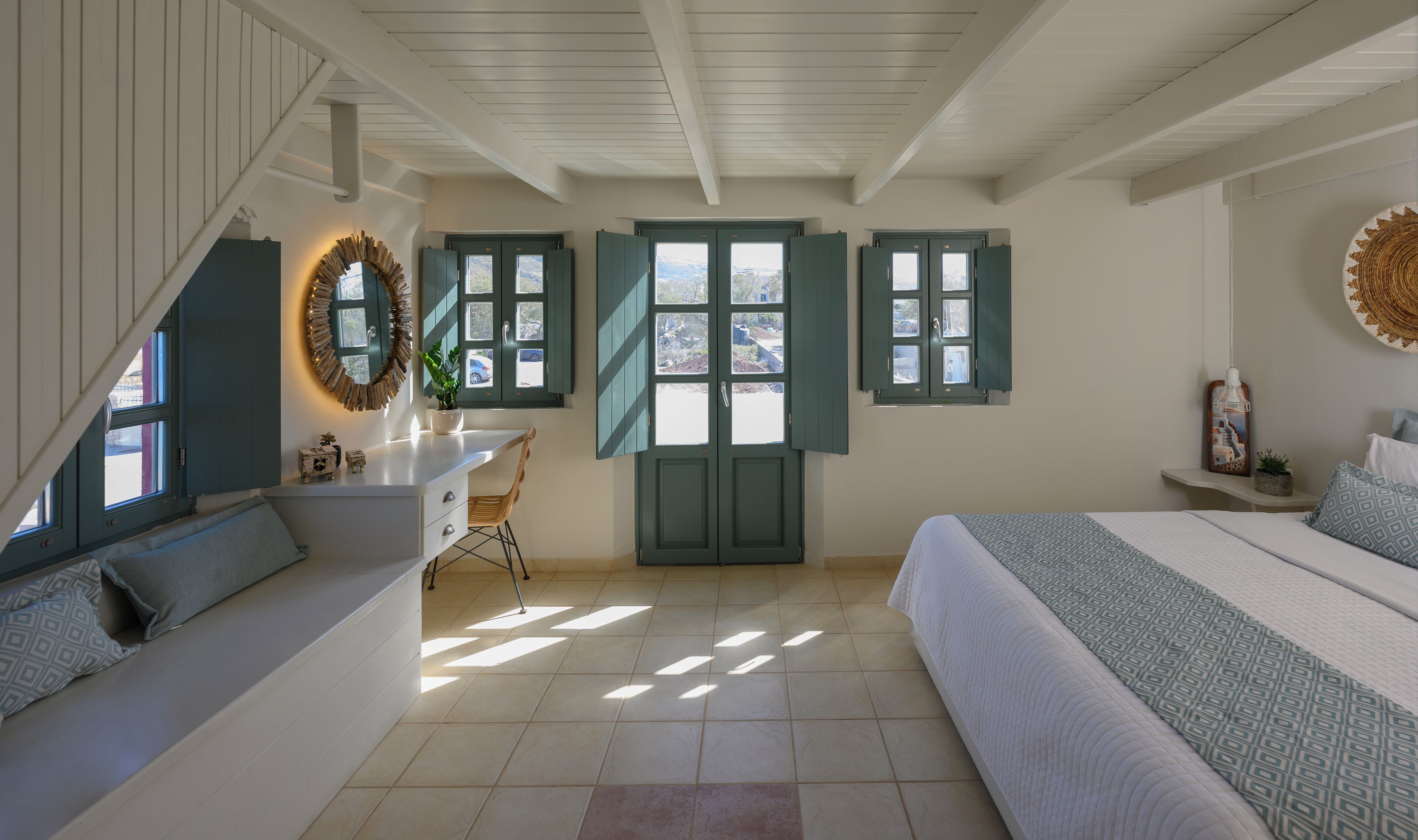 A bedroom with a bed, a desk, and a chair. There are windows with shutters, a ceiling with wooden beams, and a wall with a decorative plate.