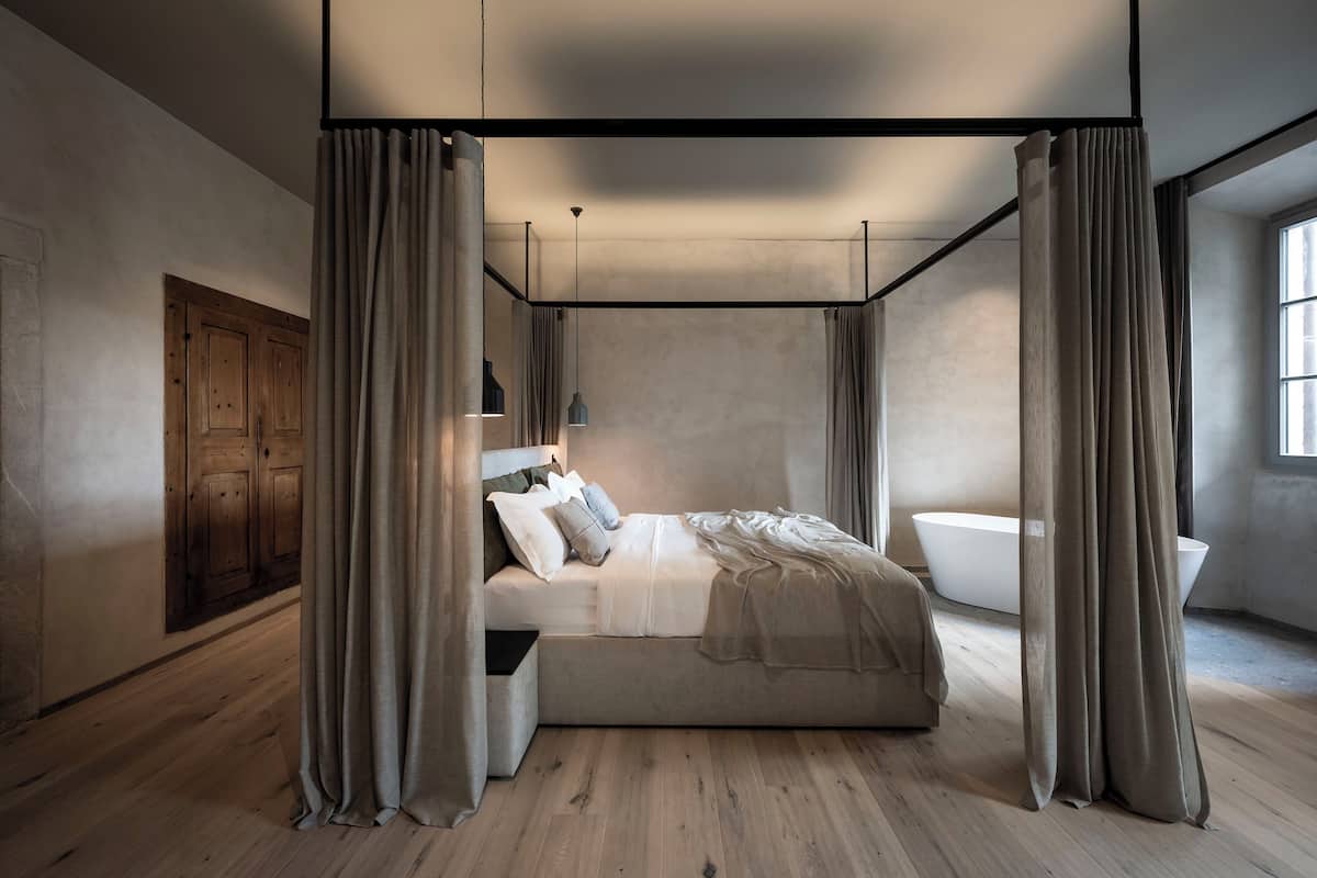 A modern bedroom with a canopy bed, wooden flooring, a large window, and a bathtub.