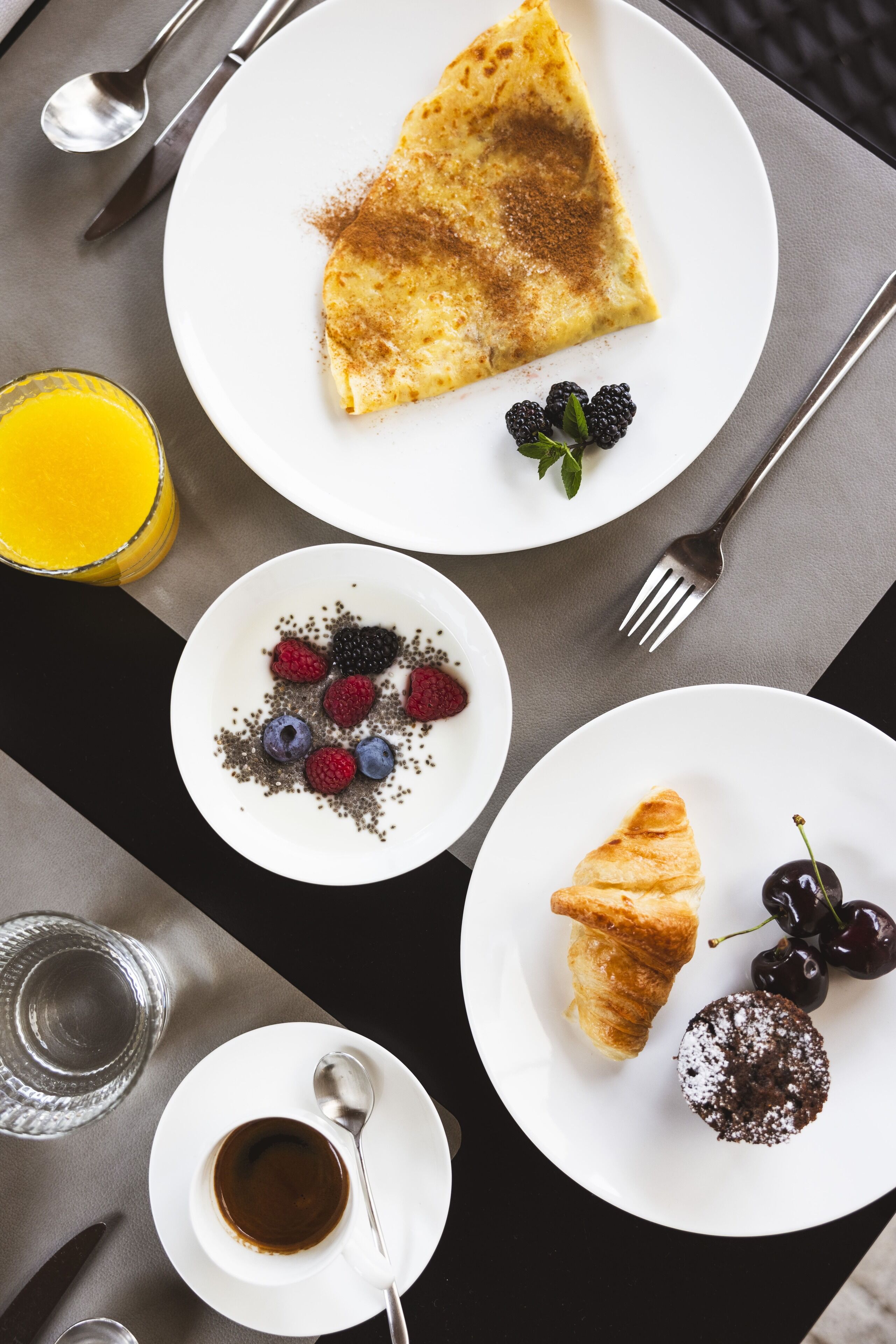 A breakfast setting with a crepe, berries, a croissant, and a muffin on plates, along with a glass of orange juice and a cup of coffee.