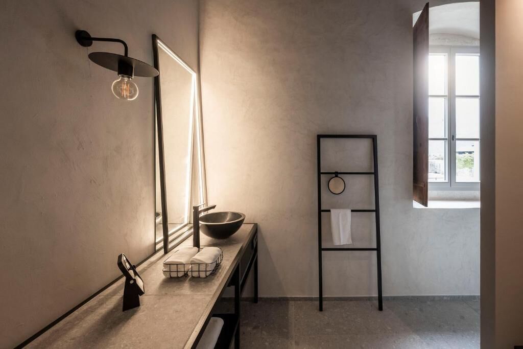 Une salle de bain minimaliste avec un grand miroir, un lavabo sur colonne, un porte-serviettes et une fenêtre laissant entrer la lumière naturelle.