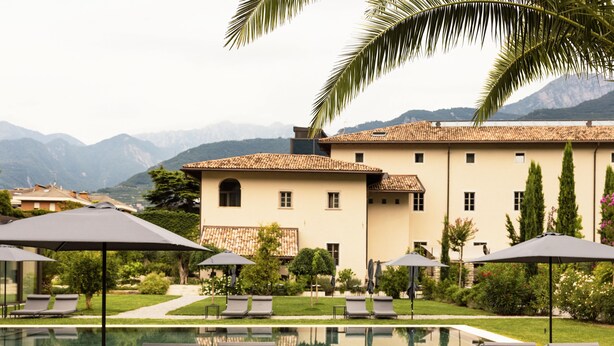 A pool area with lounge chairs and umbrellas, a building with a tiled roof, and mountains in the background. - Monastero ARX VIVENDI (Arco)