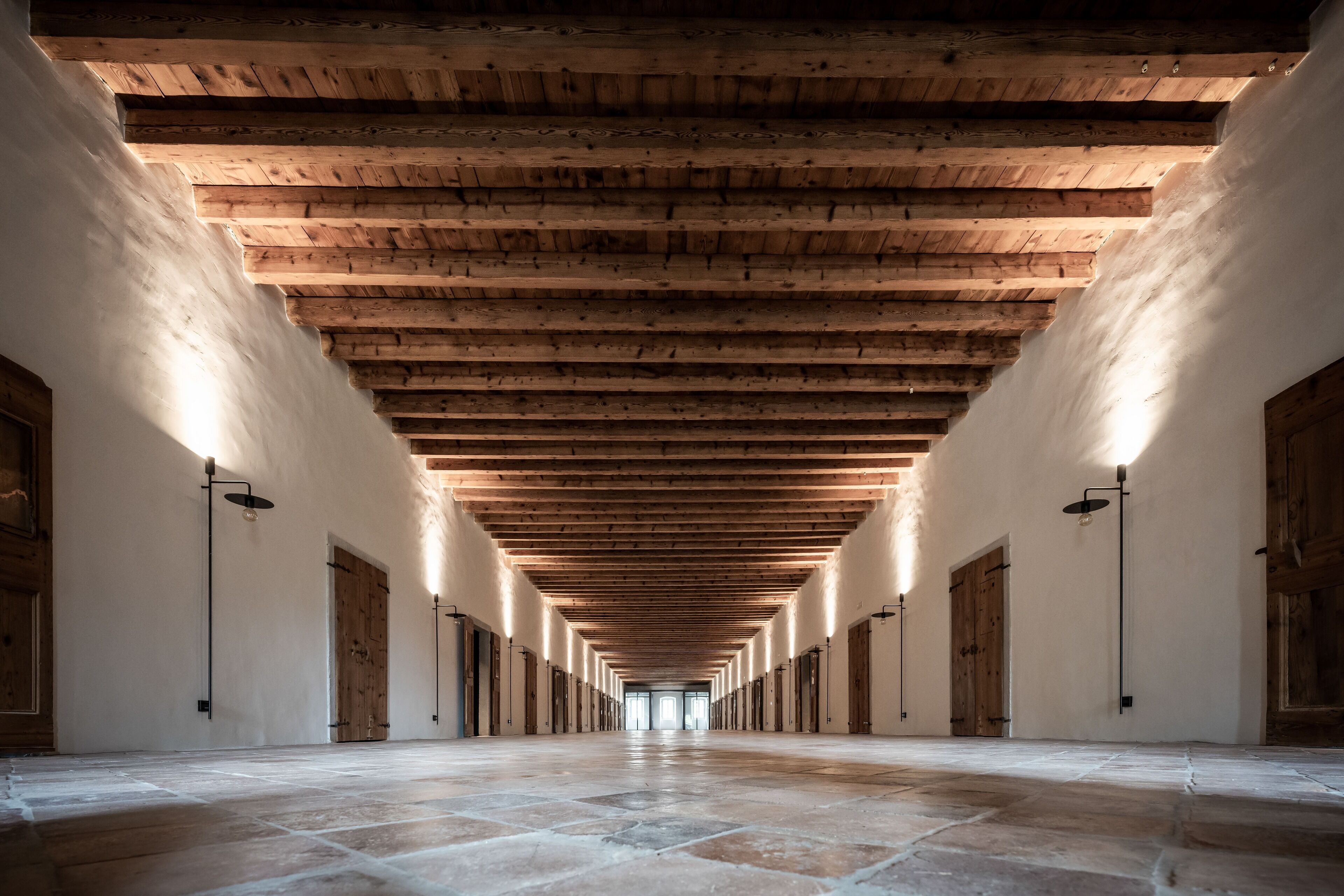 A long corridor with wooden beams and stone flooring, flanked by doors and wall-mounted lights.