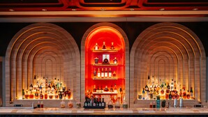 A bar with multiple shelves of liquor bottles, a central display case, and six wicker bar stools. - City of Dreams Mediterranean - Integrated Resort, Casino & Entertainment (Limassol)