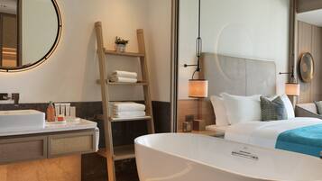 Una habitación de hotel moderna con una bañera grande, una escalera de madera, un espejo redondo y una cama con ropa blanca.