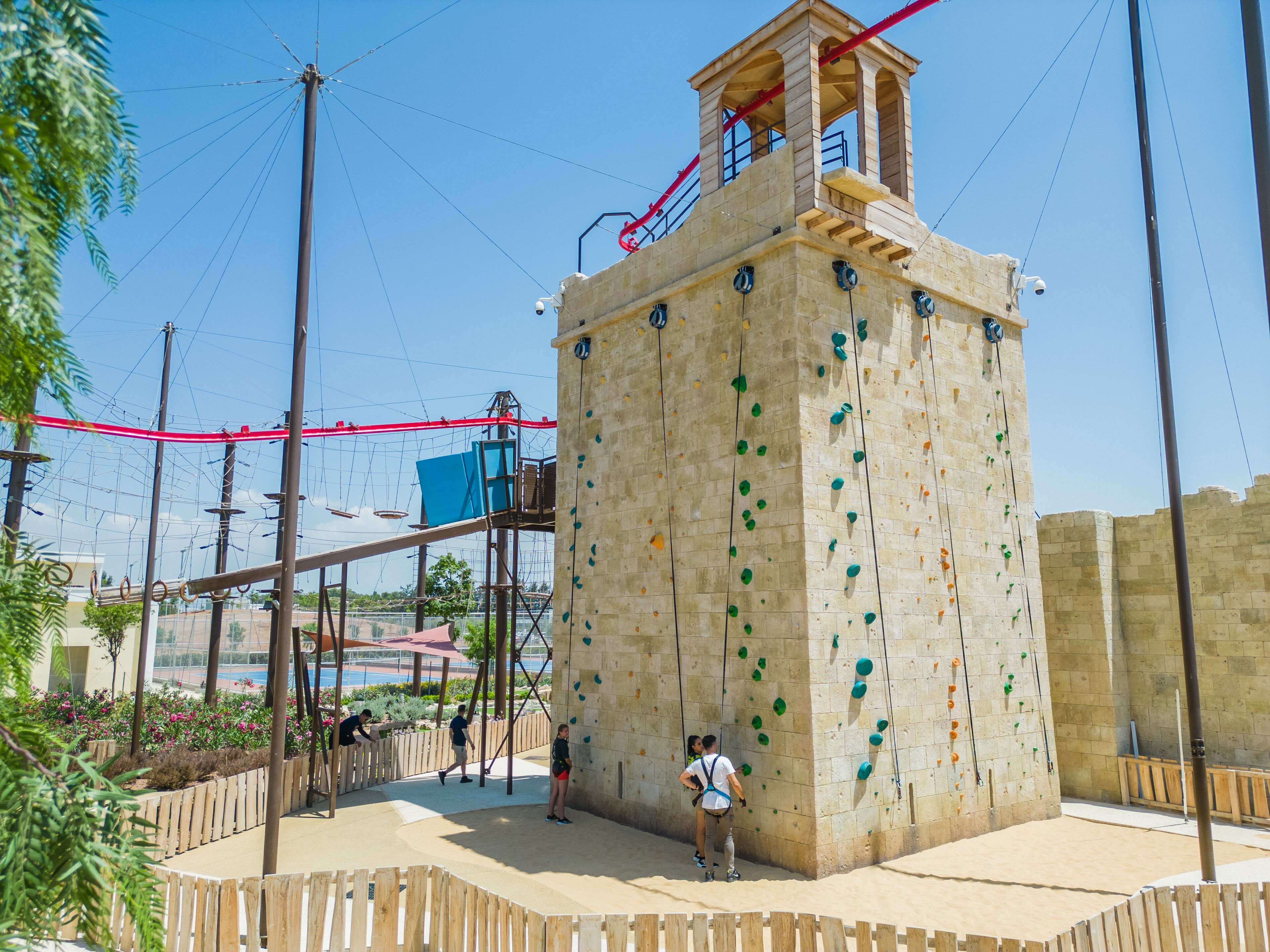 A climbing wall with a wooden observation tower, surrounded by a sandy area and climbing equipment.