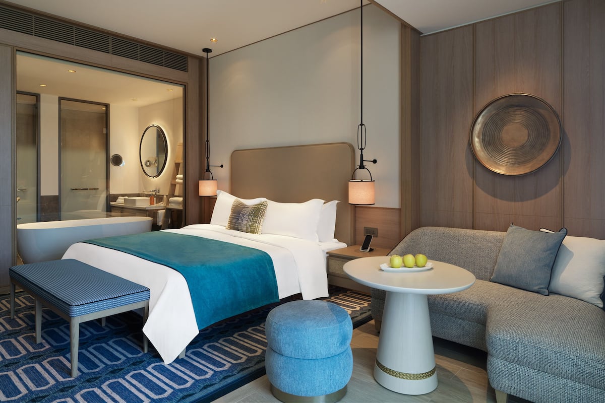 Ein modernes Hotelzimmer mit einem großen Bett, einer Couch, einem kleinen runden Tisch mit Obst und einem durch eine Glastür einsehbaren Badezimmer.