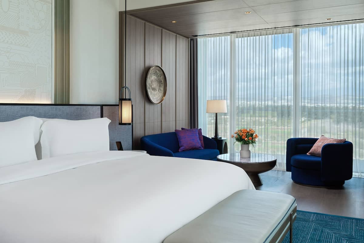 Ein Hotelzimmer mit einem großen Bett, zwei Sofas, einem kleinen Tisch mit Blumen und Blick auf die Stadt durch große Fenster.