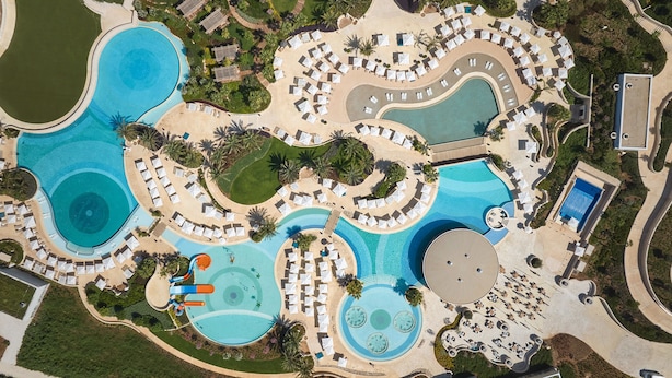 An aerial view of a resort with multiple swimming pools, sun loungers, and palm trees. - City of Dreams Mediterranean - Integrated Resort, Casino & Entertainment (Limassol)