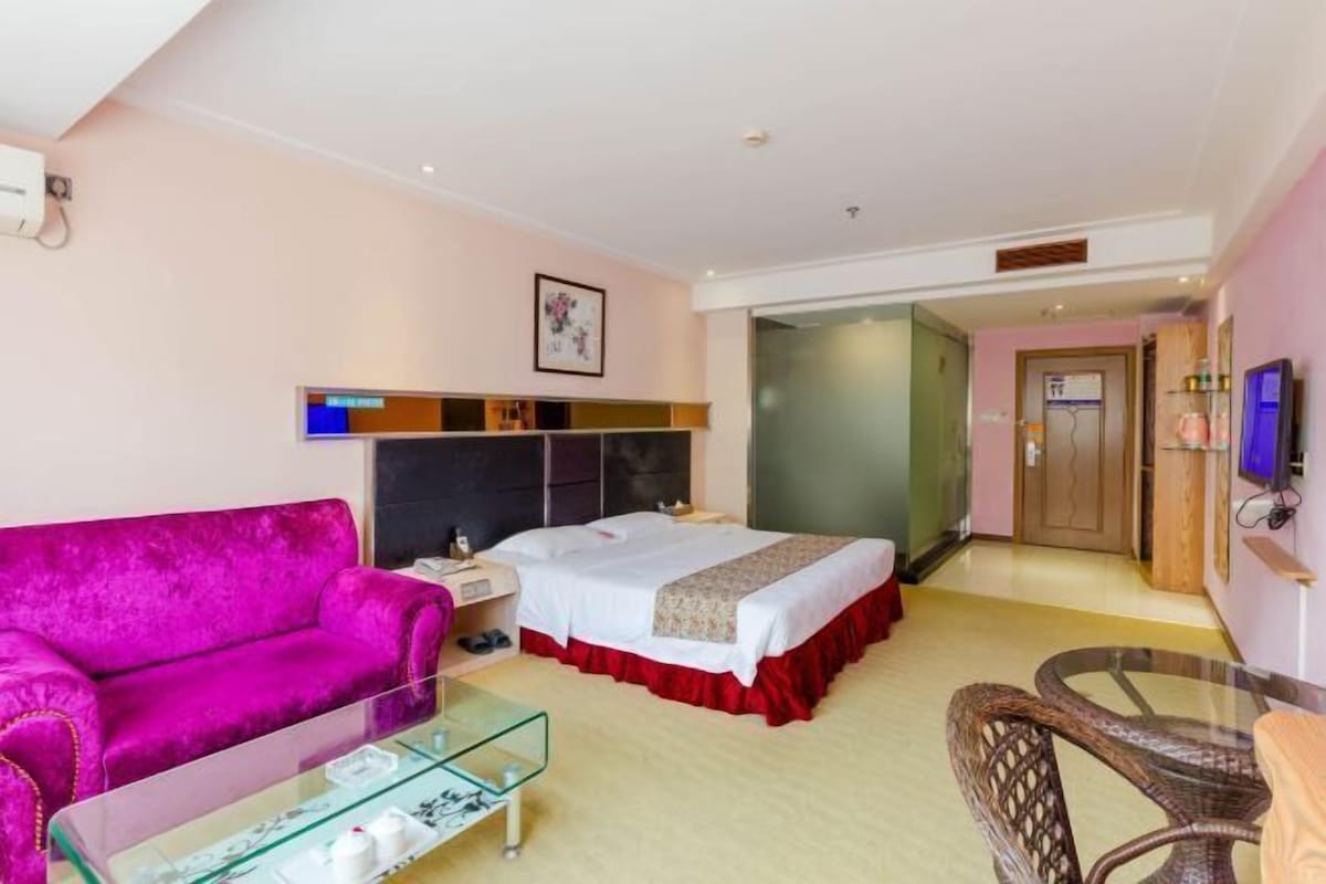 Quarto de hotel com uma cama, um sofá rosa, uma mesa de centro de vidro e uma cadeira de vime.