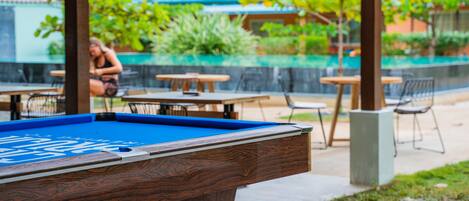 A pool table with a blue felt surface and a wooden frame. In the background, there are outdoor tables and chairs, and a person is sitting near a pool.