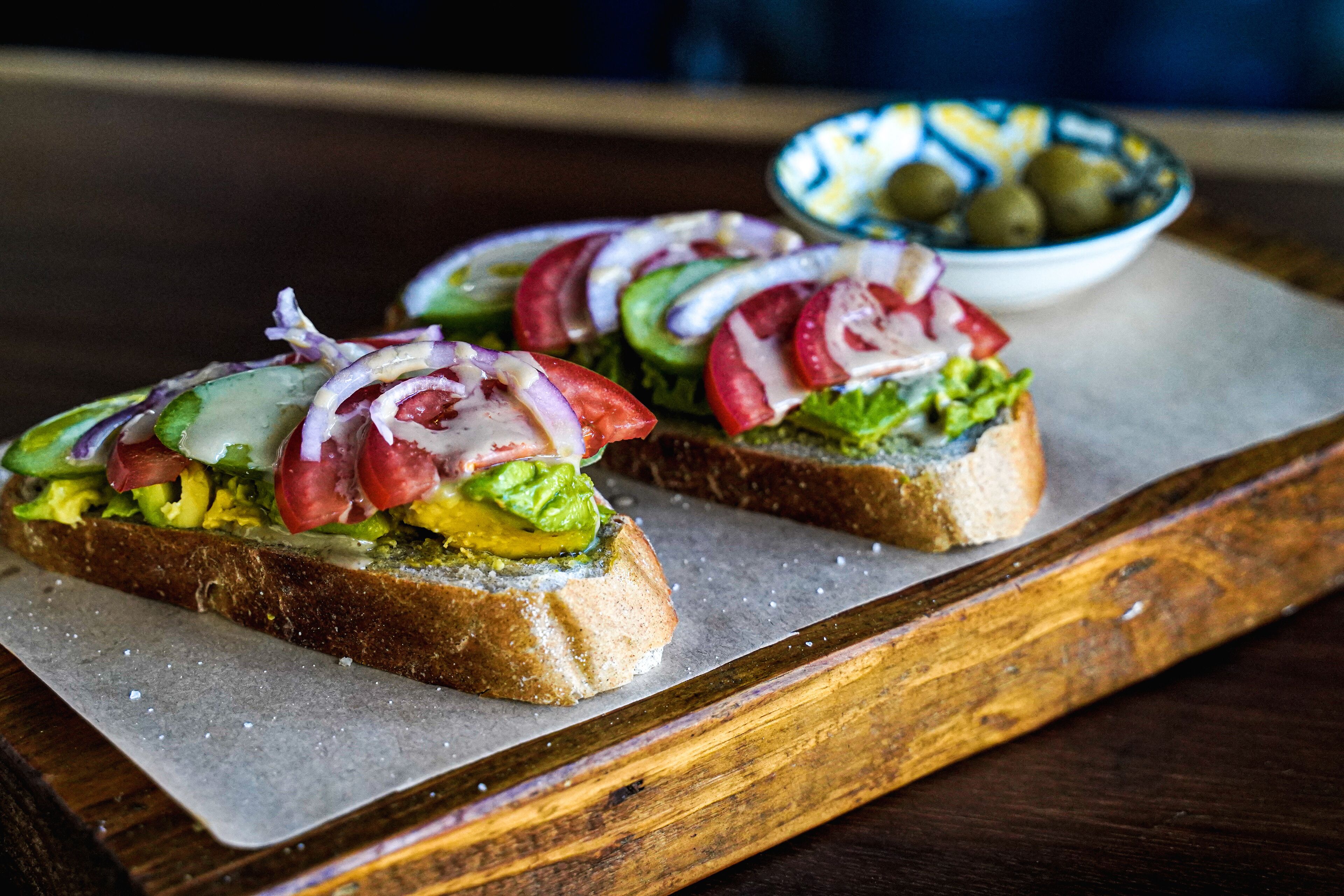 Two open-faced sandwiches on a wooden board with a bowl of olives in the background.