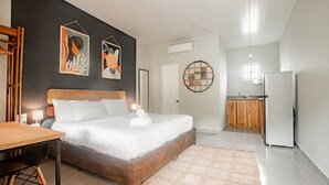 A bedroom with a wooden bed, a small table, and a kitchen area in the background. - Selina River Venao (Las Escobas del Venado)